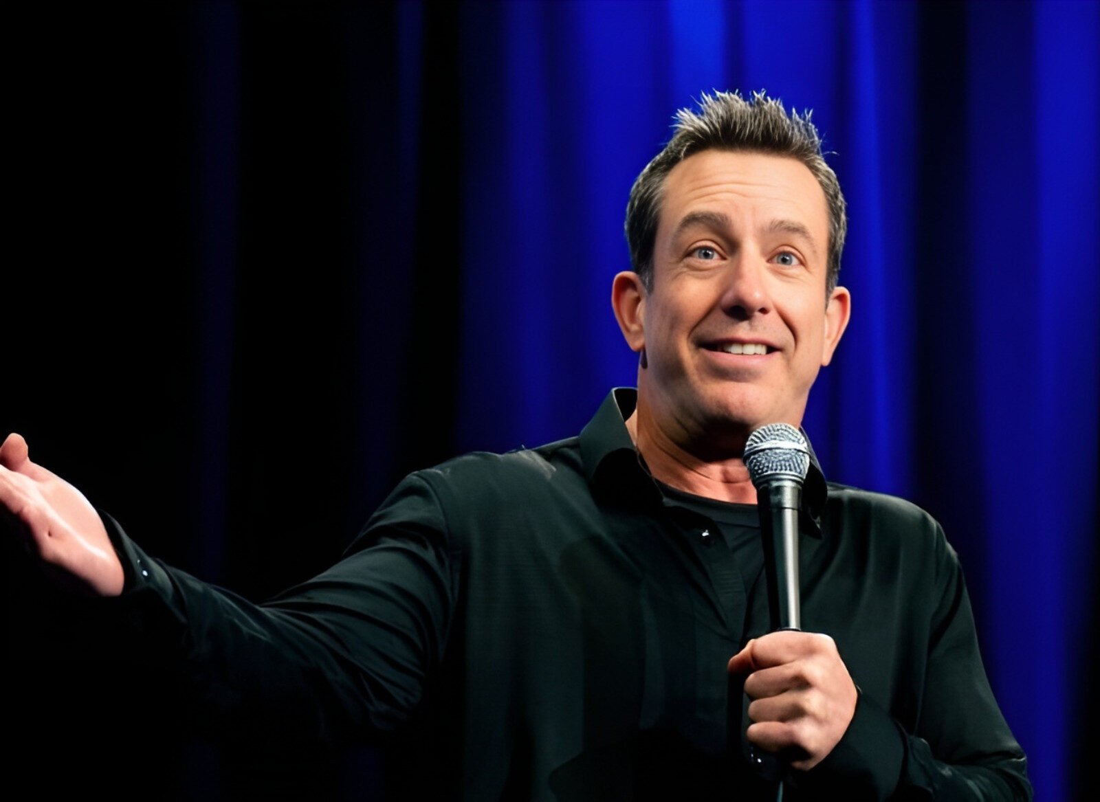 Stand-up comedian with microphone on stage, smiling during live comedy show against blue curtain backdrop