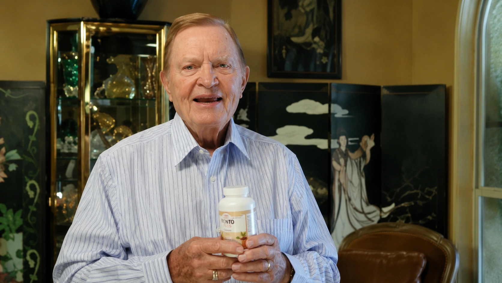 Elderly man smiling while holding a bottle of dietary supplement in a well-decorated room with decorative glass and artwork in the background.