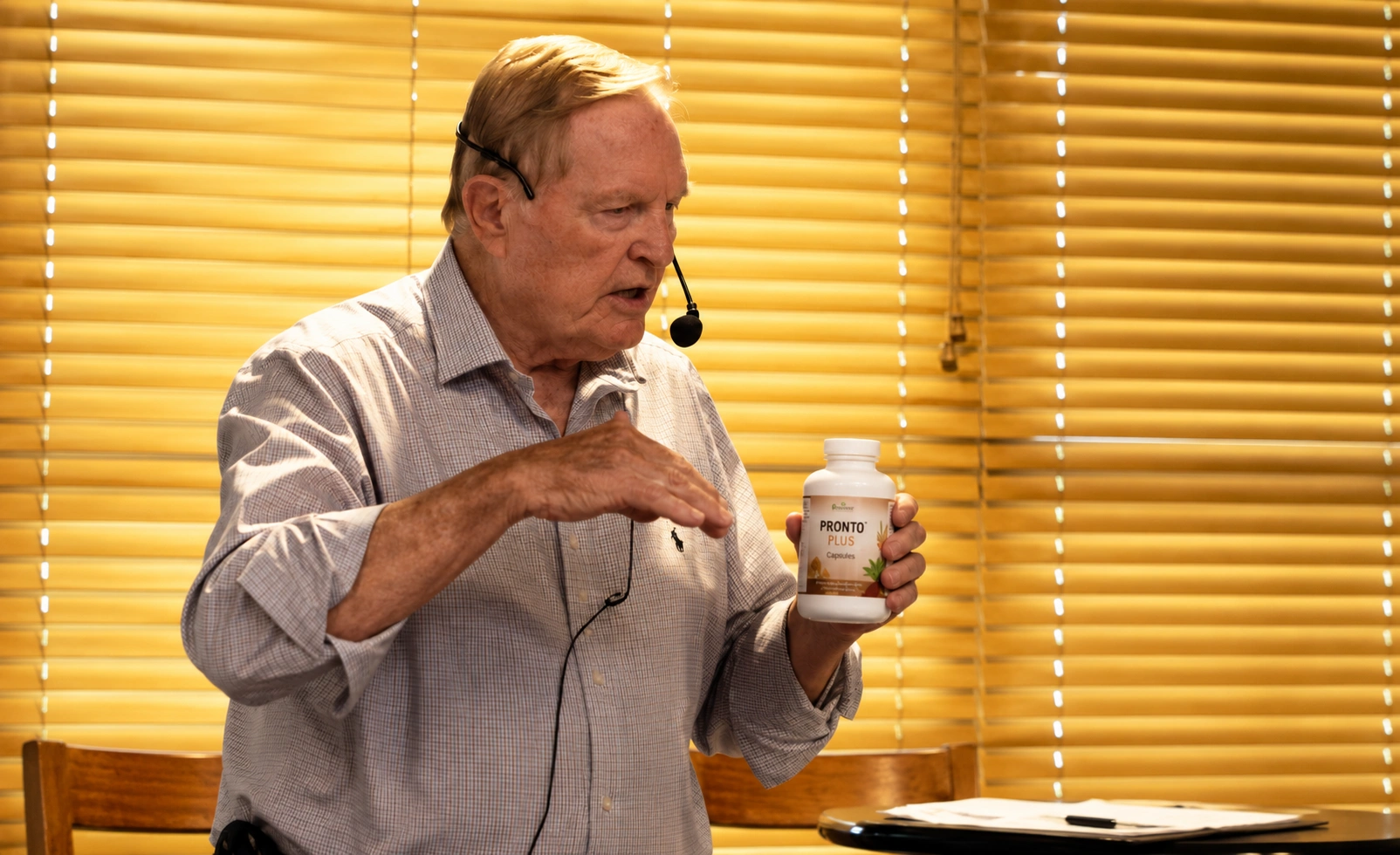 A speaker holding a bottle of Pronto Plus capsules, discussing its benefits during a presentation, with wooden blinds in the background.