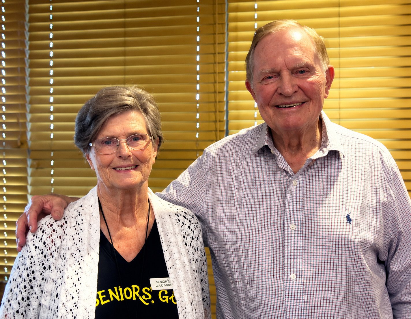 Smiling senior couple standing together indoors, with yellow blinds in the background, promoting community engagement among seniors. The woman wears glasses and a black shirt with "Seniors Go" printed, while the man is dressed in a plaid shirt.