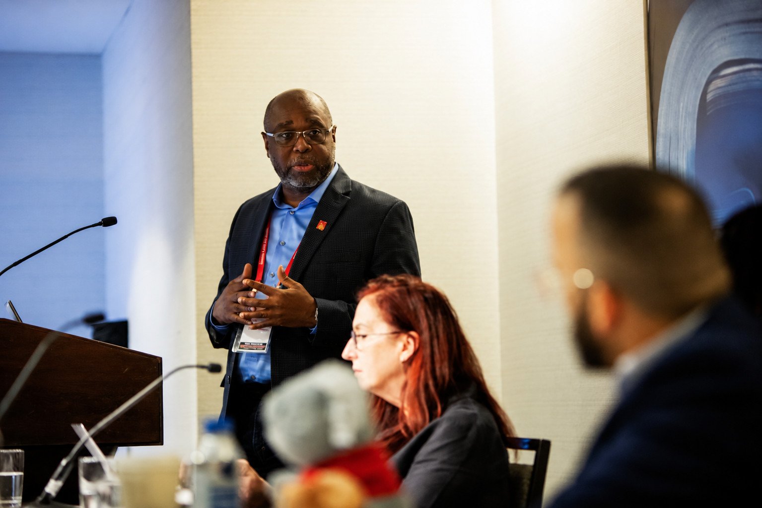 Professional speaker presenting at a conference, engaging with an audience seated at a table, showcasing a collaborative discussion in a business setting.