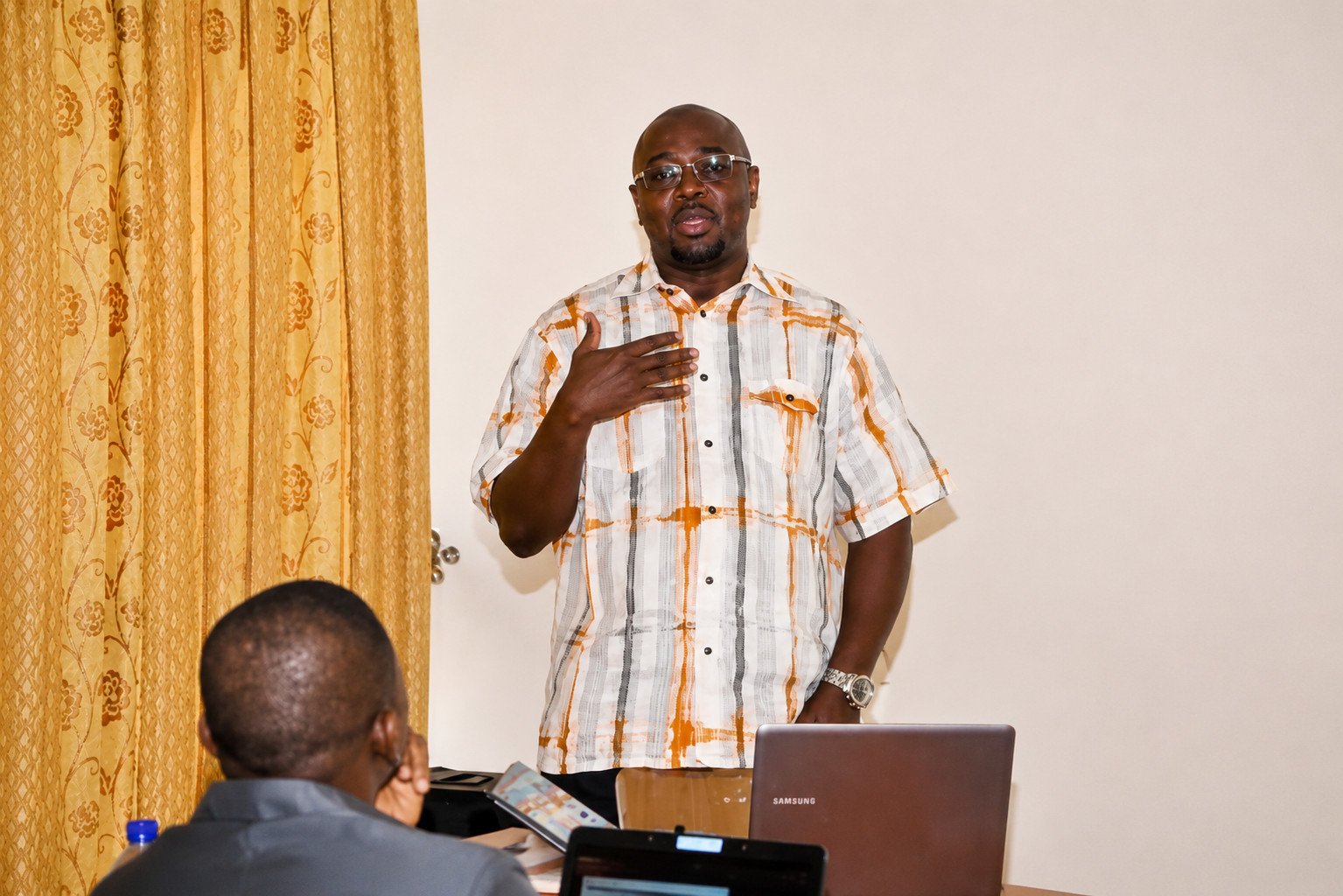 A man in a patterned shirt delivers a presentation in a conference room, while seated individuals focus on their laptops and notes. The setting features yellow curtains and a professional atmosphere.