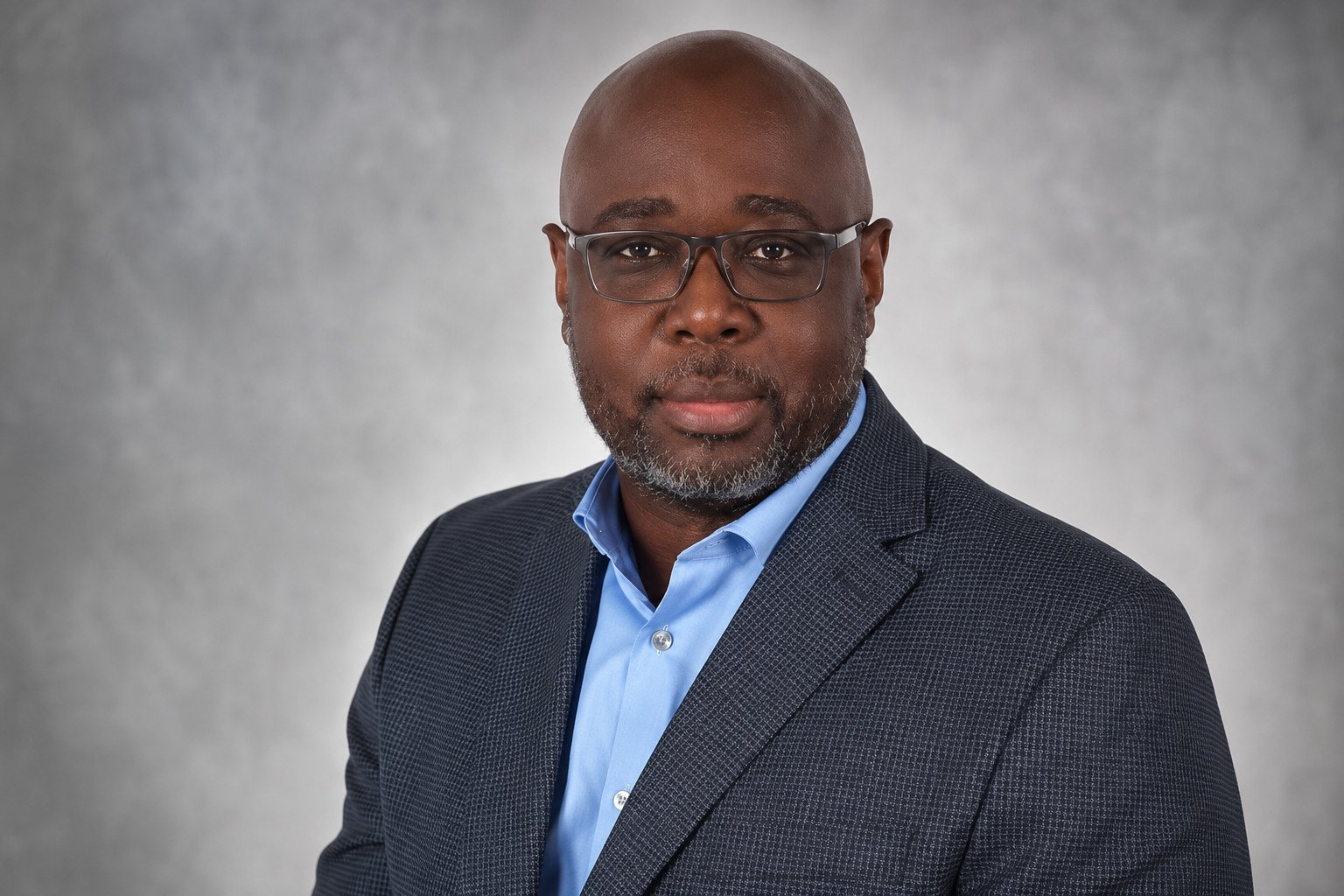 Professional headshot of a middle-aged man with a bald head and glasses, wearing a dark blazer over a light blue shirt, set against a neutral gray background.