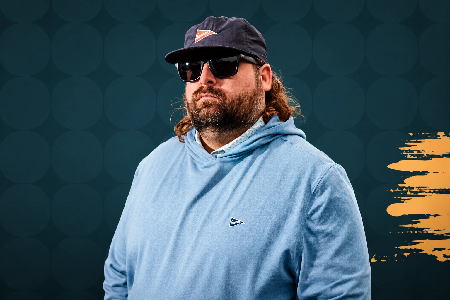Man wearing sunglasses and a navy cap, dressed in a light blue hoodie, posing against a dark teal background with circular patterns. The image conveys a casual and stylish vibe, suitable for fashion or lifestyle content.