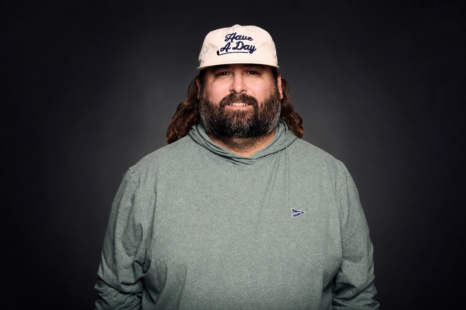 Fat Perez Net Worth – Portrait of Fat Perez with long hair and a beard, wearing a green hoodie and a beige "Have A Day" cap against a dark background.