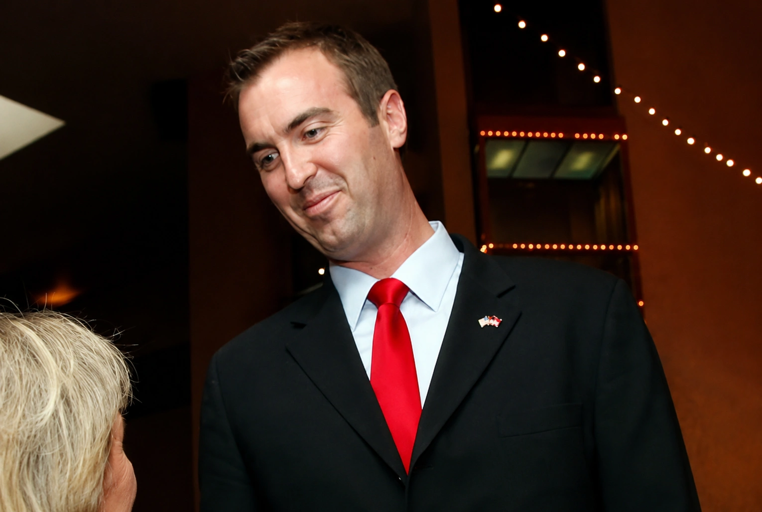 Jesse Kelly Net Worth – Tall man in a black suit with a red tie, smiling and interacting with a woman at a formal event, with decorative lighting in the background.
