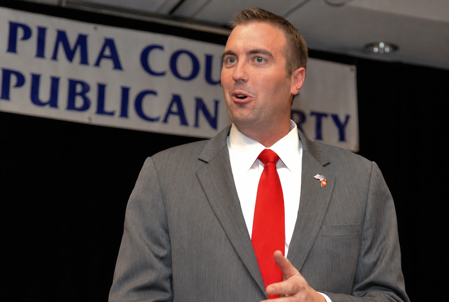 Man in a gray suit and red tie speaking at a Pima County Republican Party event, with a banner in the background.