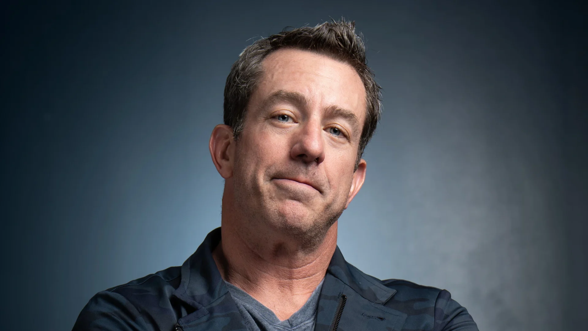Jamie Lissow wearing a blue shirt under a black jacket stands confidently, showcasing a stylish and professional appearance.